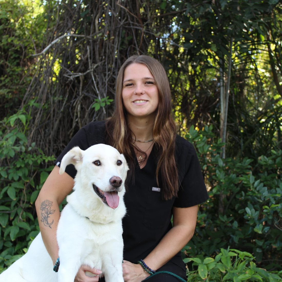 Woman in black scrubs with a white dog in front of greenery